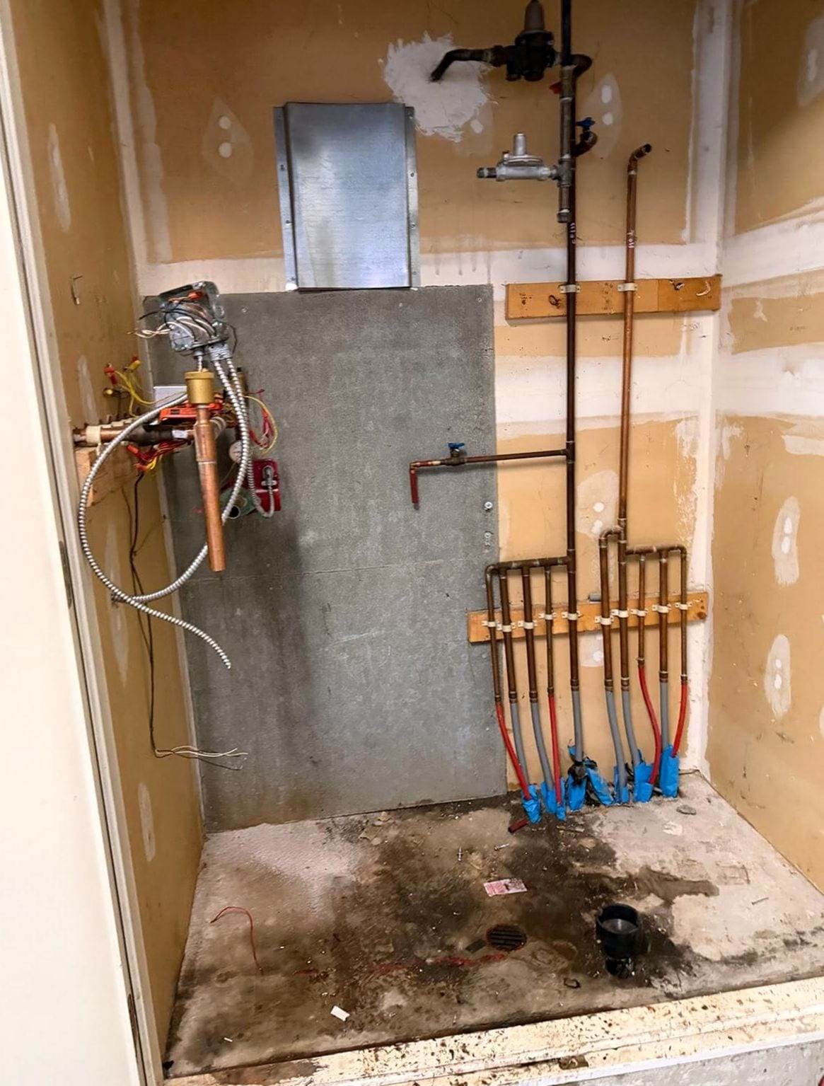 Utility closet with the old heating unit removed, leaving the gas line and regulator, copper piping and a row of red and gray PEX heating loops coming up through the floor