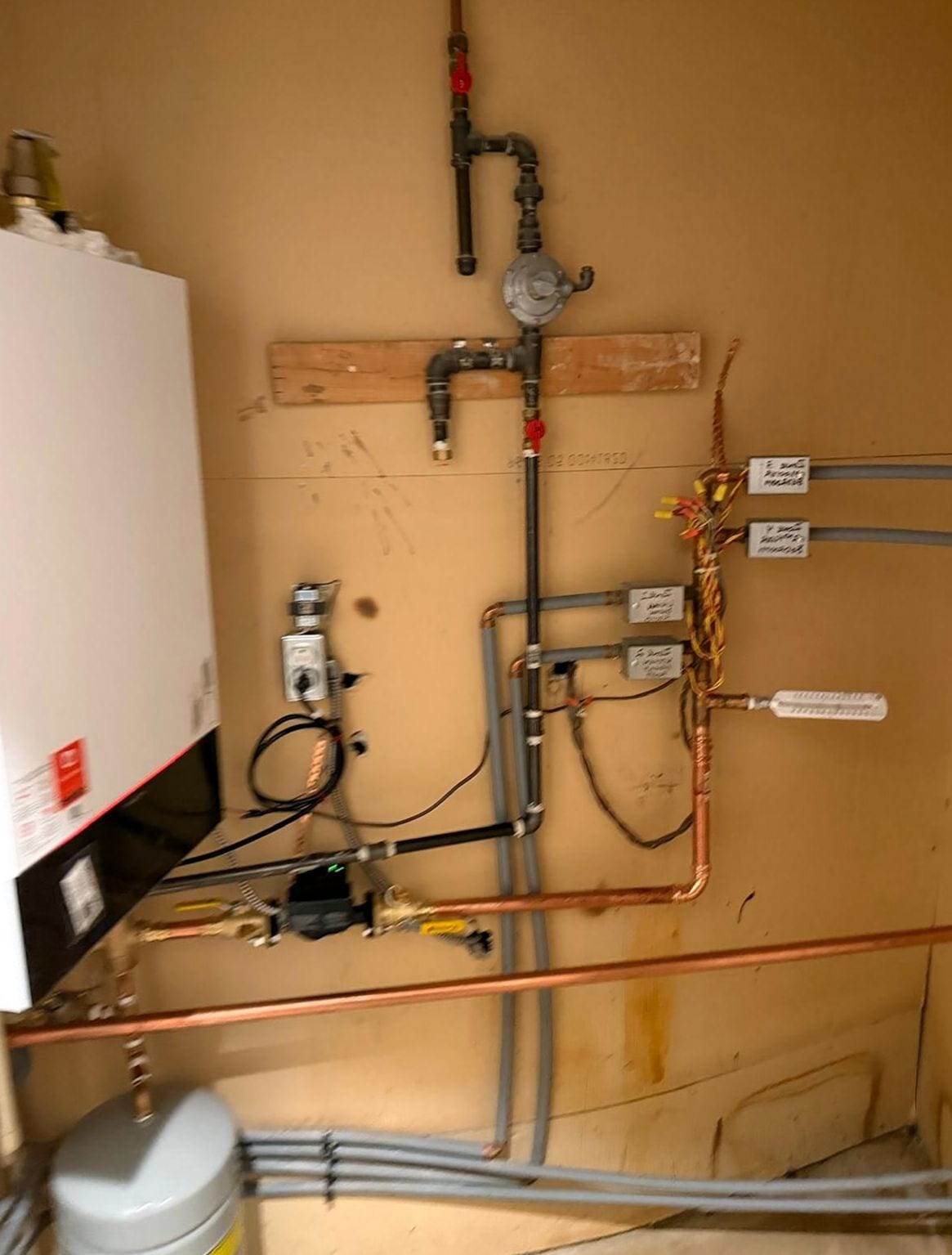Wall-hung gas boiler with its near-boiler piping: black gas pipe and regulator, a circulator pump between yellow-handled valves, copper lines, labeled zone wiring and an expansion tank