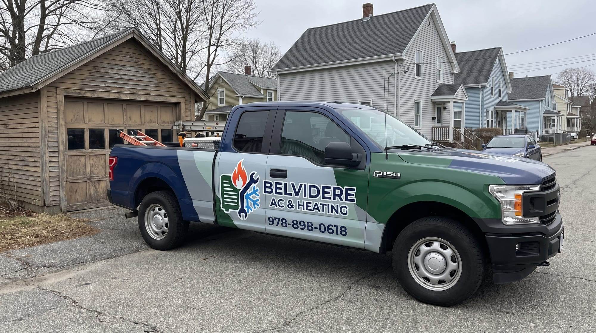 Belvidere AC & Heating pickup truck wrapped in navy blue, light gray and forest green, with the company logo and the phone number 978-898-0618 on the door, parked on a street of older clapboard houses